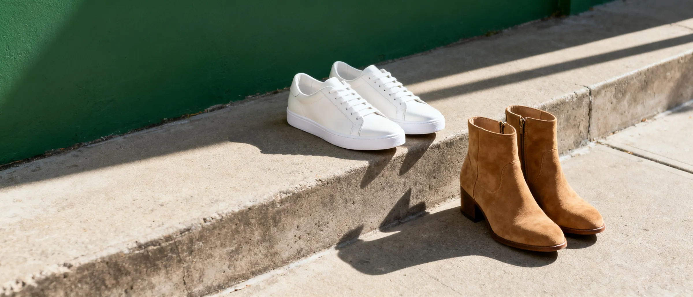 A pair of white canvas sneakers and a pair of tan suede boots on a sunlit concrete step